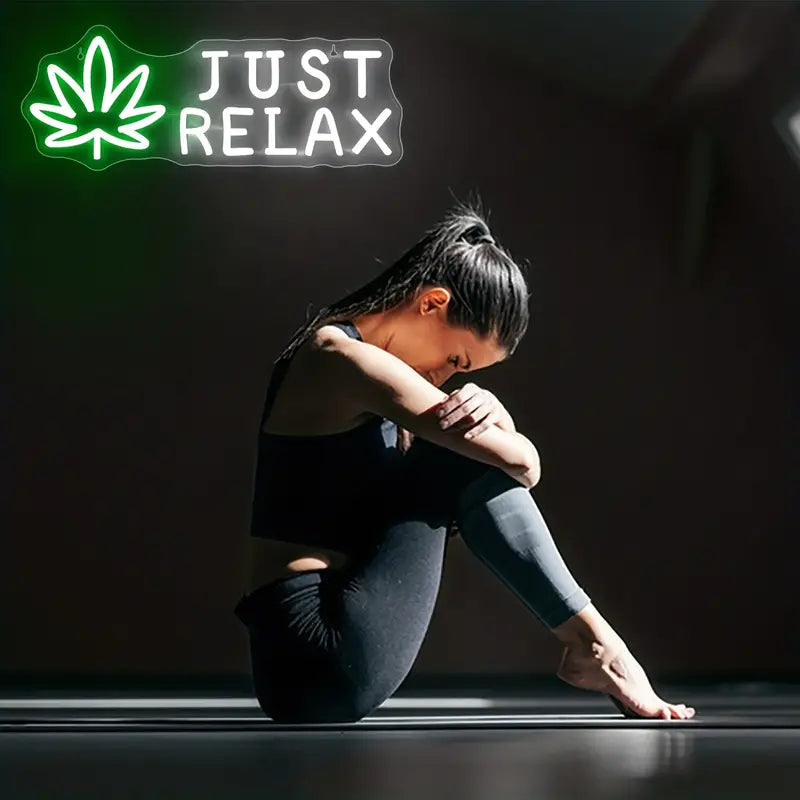 Woman embracing knees under neon "Just Relax" sign, leaf design.