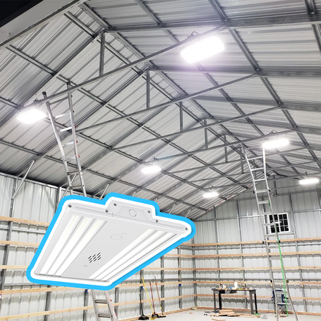 A close-up of a white LED high bay light fixture, with a background showing several of the same lights installed and illuminating a large metal workshop with a high steel-truss ceiling
