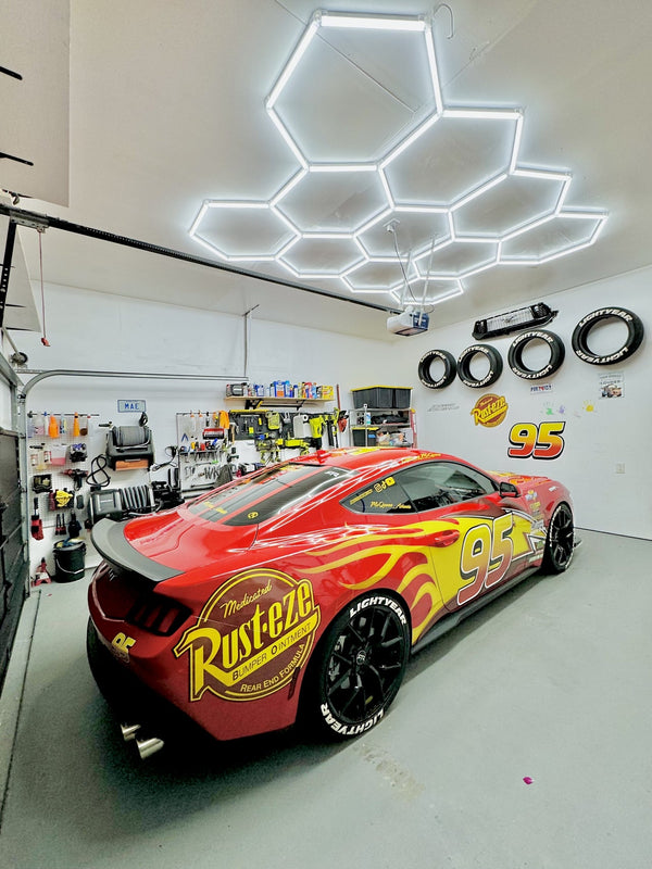 Residential garage workshop with modular hexagon LED ceiling panels delivering bright, uniform task lighting over a cluttered workbench, tool cabinets, and vehicle frames; visible junction/cable suggests DIY install—relevant for garage shop lighting, hexagon light kits, and installation guides.