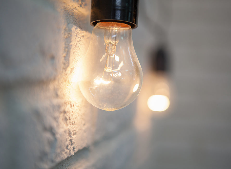 A close-up of a lit incandescent light bulb in a black socket, casting a warm glow against a textured white brick wall. A second bulb is visible but out of focus in the background.