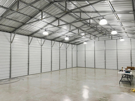 A spacious and empty steel-framed warehouse with white panel walls, a concrete floor, and a row of UFO high bay lights.