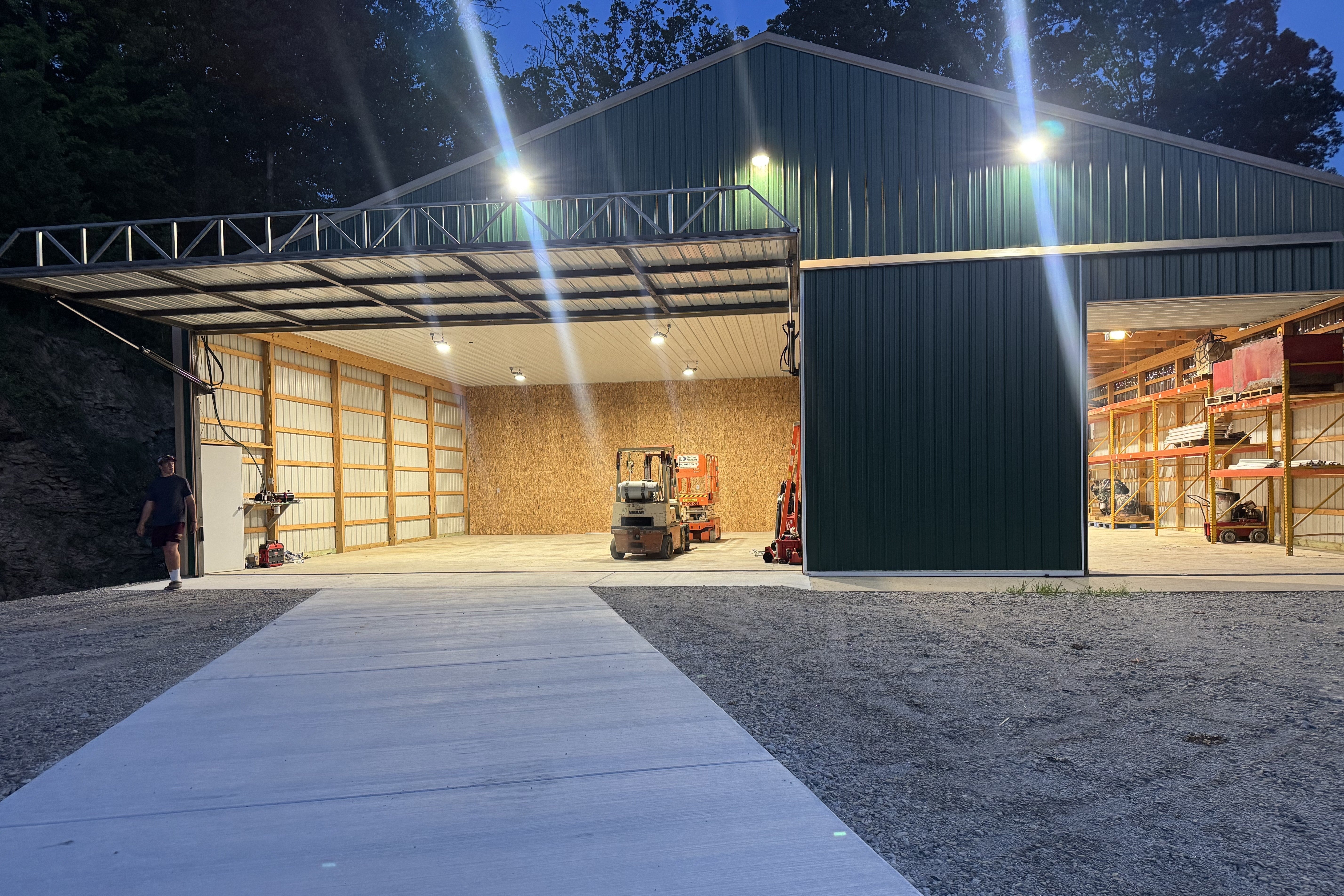 A large green metal warehouse at dusk, brightly illuminated by LED lights. The large bay doors are open, revealing a well-lit interior with a forklift and a separate storage area with shelving.