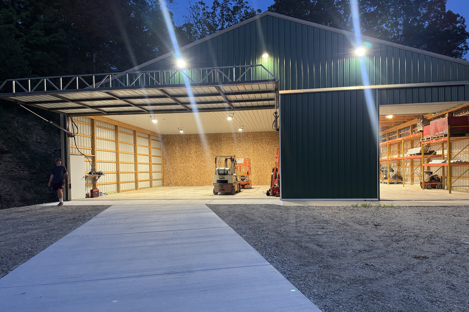 A large green metal warehouse at dusk, brightly illuminated by LED lights. The large bay doors are open, revealing a well-lit interior with a forklift and a separate storage area with shelving.