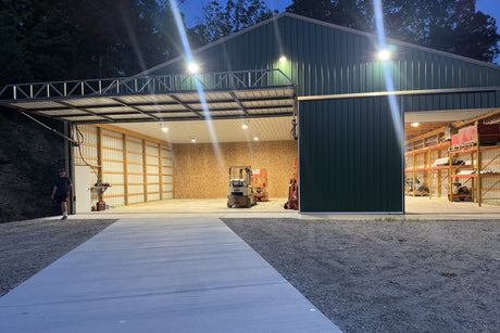 A large green metal warehouse at dusk, brightly illuminated by LED lights. The large bay doors are open, revealing a well-lit interior with a forklift and a separate storage area with shelving.