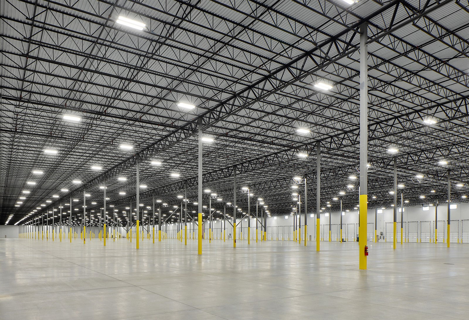 Spacious empty warehouse interior featuring numerous linear high-bay lights evenly installed on the ceiling, illuminating the area with bright, uniform light, supported by metal columns with yellow bases.