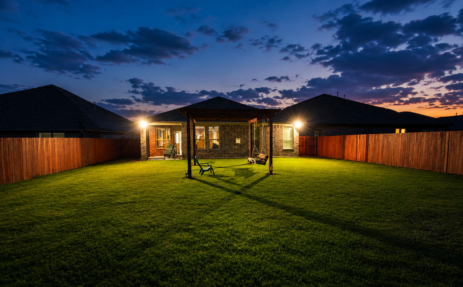 Symmetrical outdoor flood lights illuminate a spacious fenced backyard and wooden pergola under a dramatic sunset sky, enhancing both aesthetic appeal and nighttime property safety.