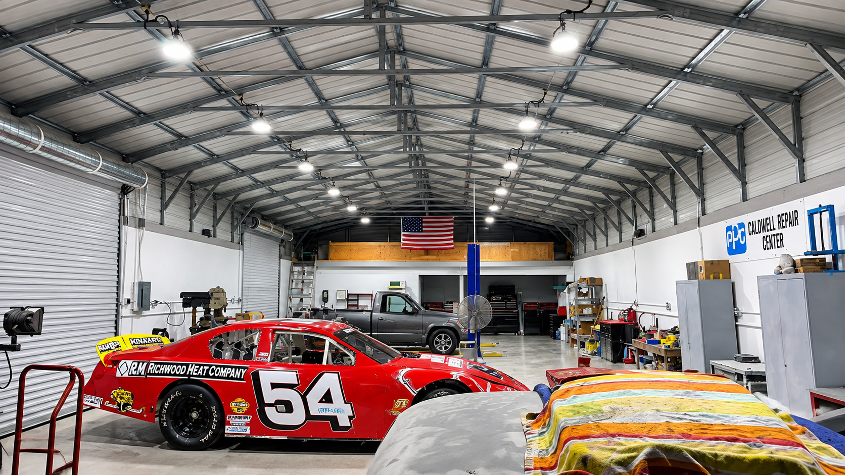 Round high bay lights suspended from the ceiling of a race car workshop above a red #54 race car and a pickup truck