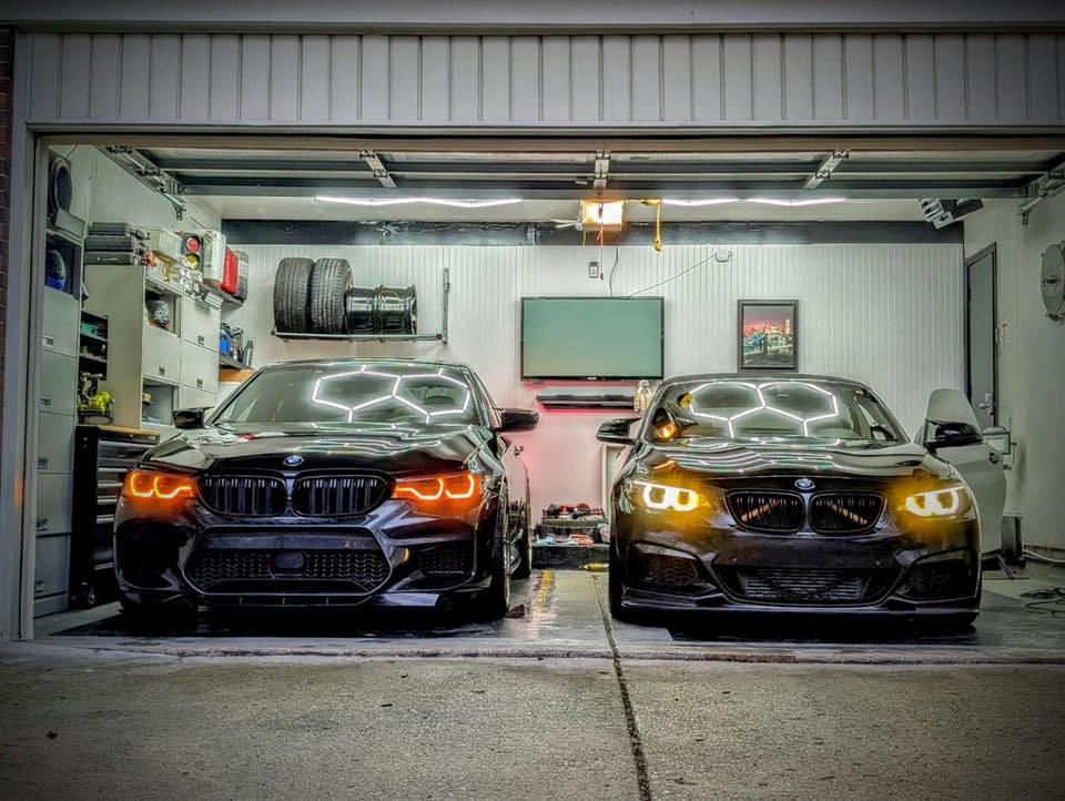Hexagon LED garage lights perfectly reflect off the glossy hoods and windshields of two black luxury cars parked side-by-side, demonstrating excellent illumination and shadow reduction in the workspace.