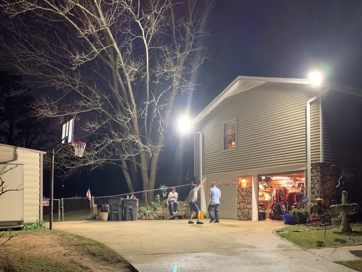 A night scene of a residential home with a flood light illuminating the outdoor area, basketball hoop, and people gathered around.