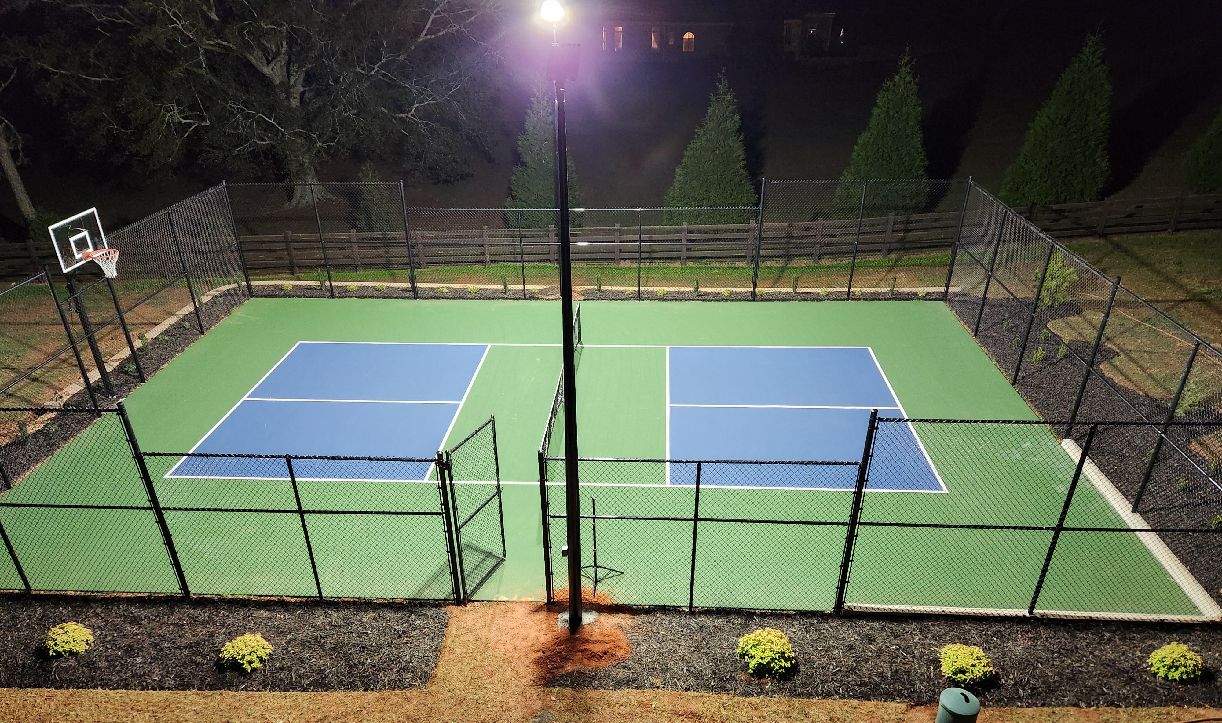 Overhead view of a fenced pickleball court at night lit by one tall flood light above green and blue courts