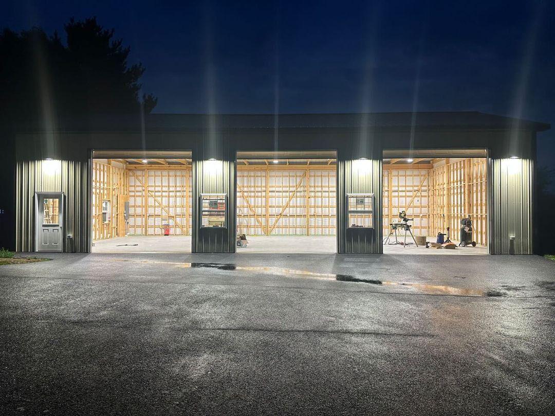 Large outdoor garage lit by powerful flood lights at night, showing broad coverage across open bays, work areas, and the wet paved approach.