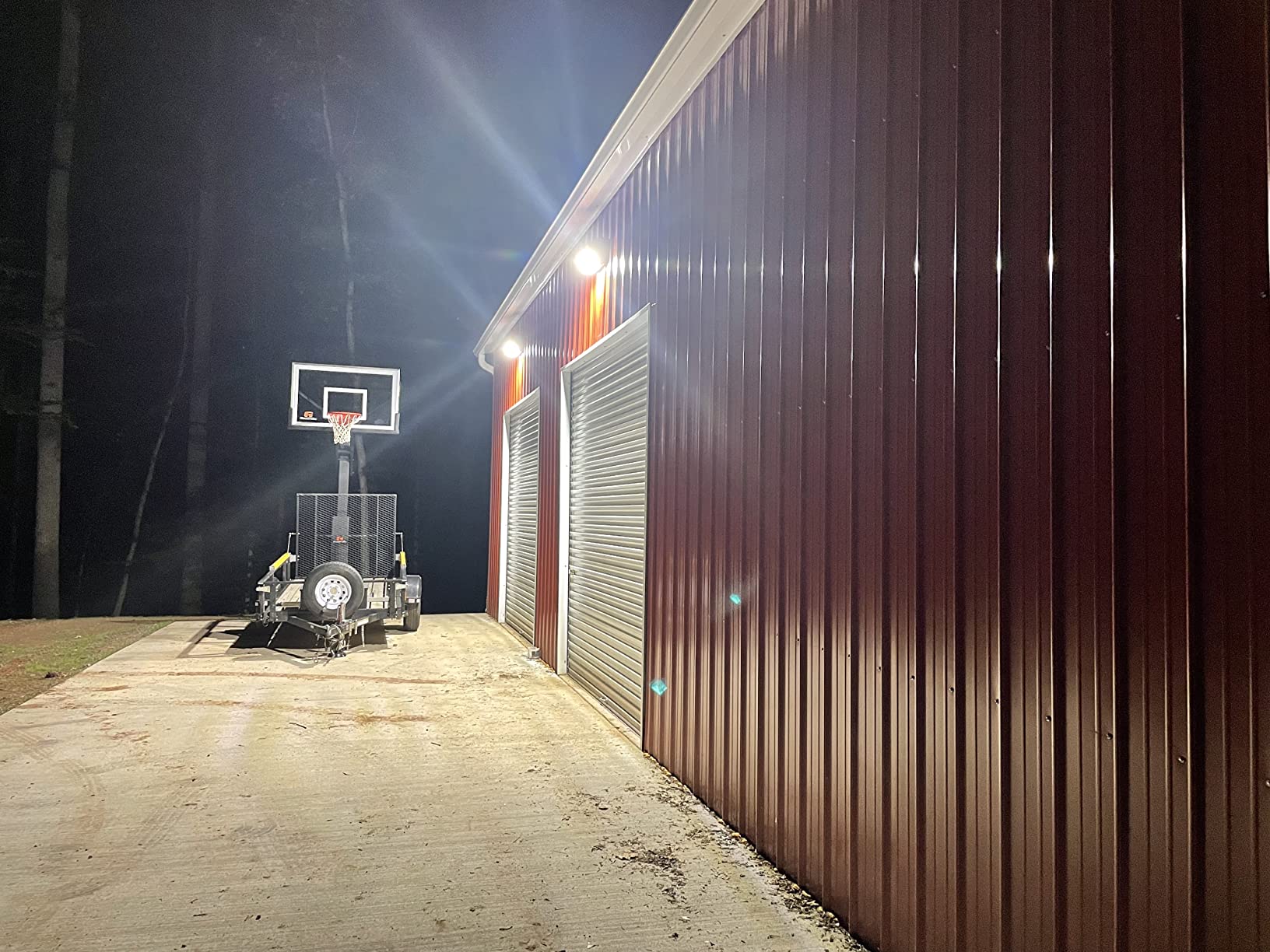 A portable basketball hoop on a concrete driveway next to a red metal building at night.