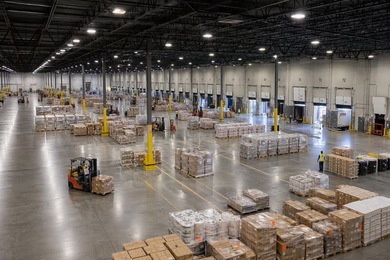 Open warehouse staging floor with evenly spaced overhead high-bay fixtures, pallets, forklifts, and a visible dock area