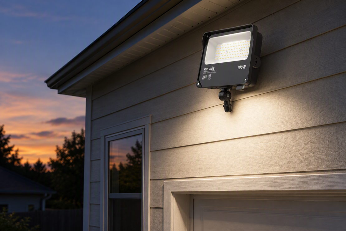 Outdoor LED floodlight mounted near a building at dusk, illuminating a nearby wall that reflects light toward the fixture’s sensor