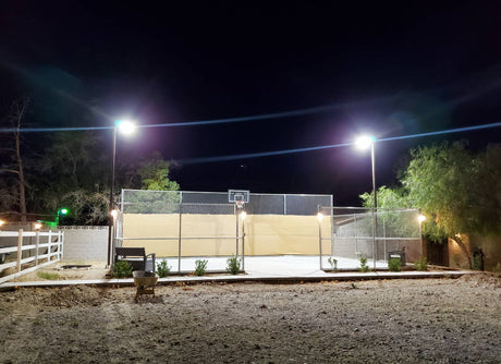 A basketball court at night lit by bright Parking Lot Lights, surrounded by fencing and desert vegetation.
