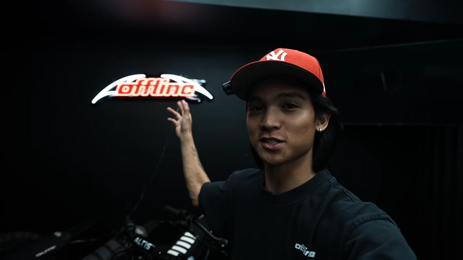 A young man wearing a red NY baseball cap and a black t-shirt gestures behind him toward a glowing "offline" LED neon sign mounted on a dark wall.