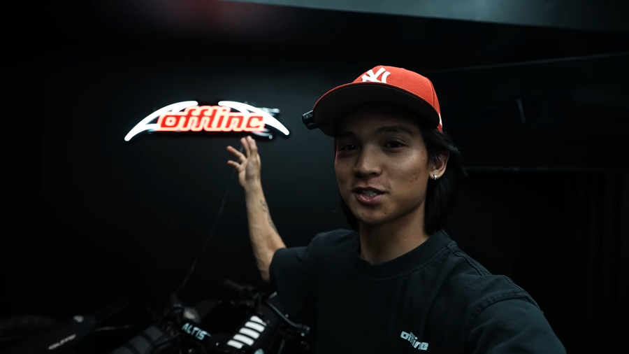 A young man wearing a red NY baseball cap and a black t-shirt gestures behind him toward a glowing "offline" LED neon sign mounted on a dark wall.