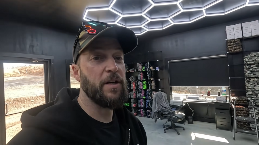 A bearded man in a black hoodie and cap speaks to the camera inside a finished office space featuring hexagonal ceiling lights, dark walls, a desk, and merchandise shelving.