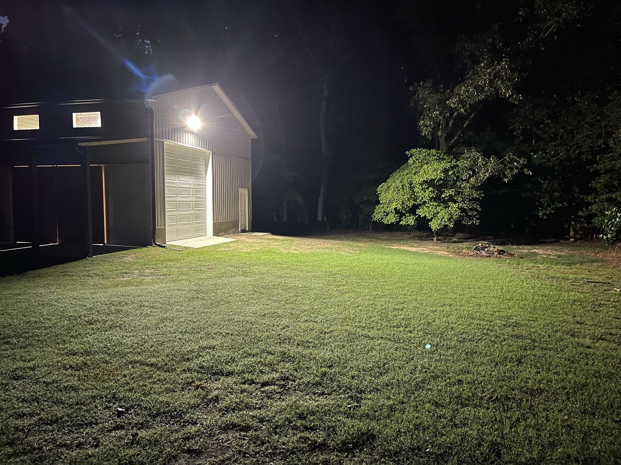 A bright outdoor LED floodlight attached to a large metal barn successfully illuminates the surrounding dark grassy yard.