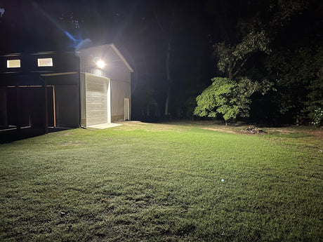 A bright outdoor LED floodlight attached to a large metal barn successfully illuminates the surrounding dark grassy yard.