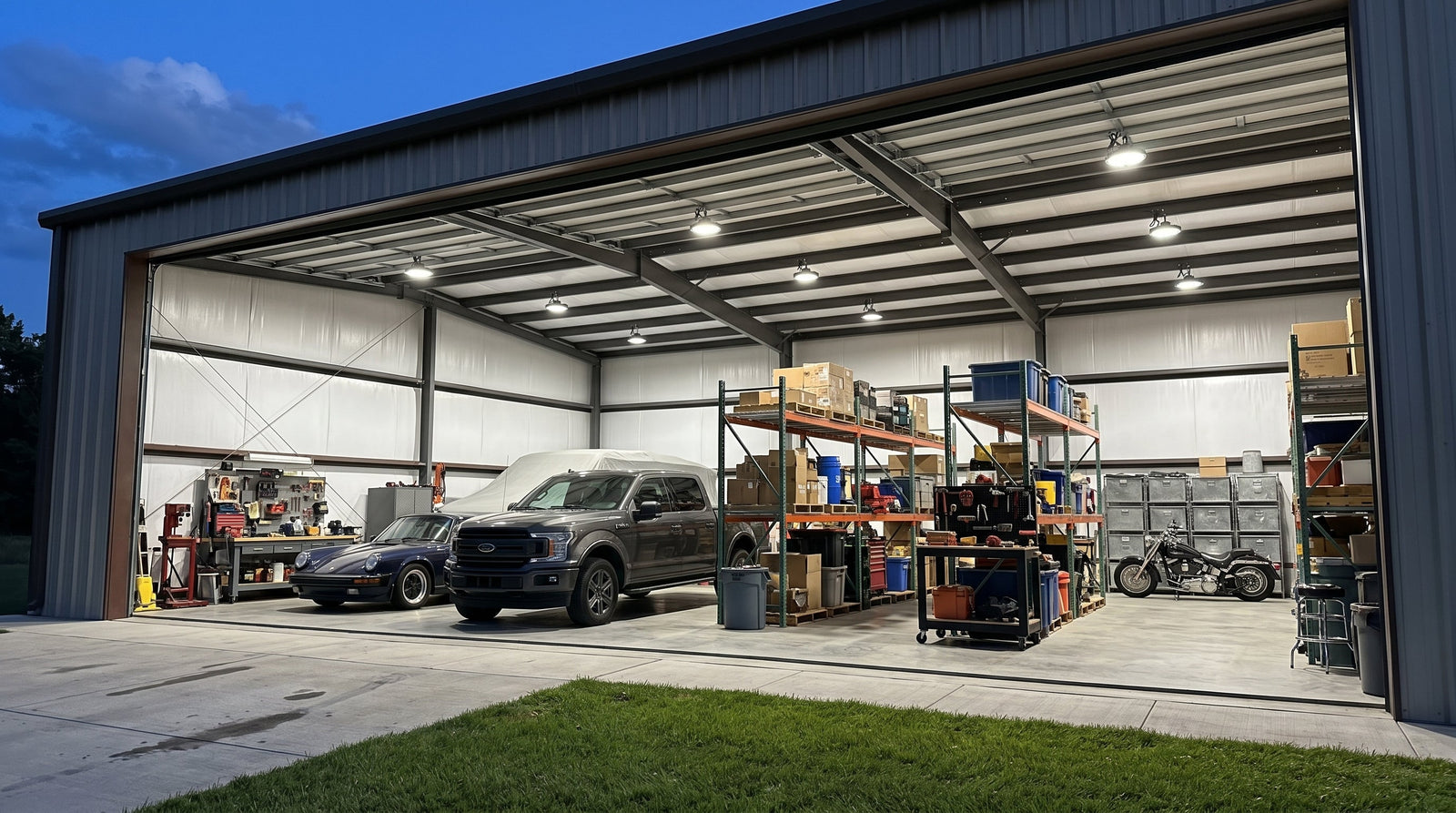 Wide view of an open commercial garage at dusk, displaying an organized storage rack, pickup truck, sports car, and motorcycle under a grid of brilliant LED lights.