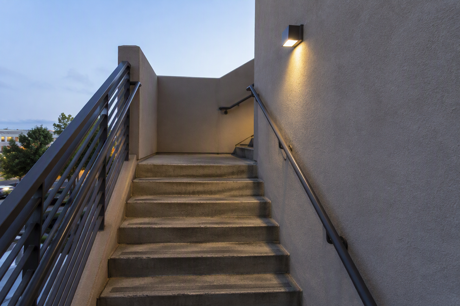 Commercial outdoor stairs at dusk, with one wall light beside a turning landing where a wall partly screens the next flight.