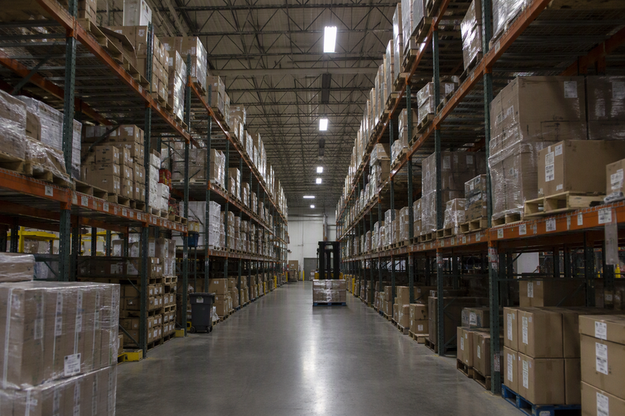 A warehouse aisle with a row of suspended linear high-bay lights; some fixtures are lit while others in the same row are dark, showing a partial visible pattern.