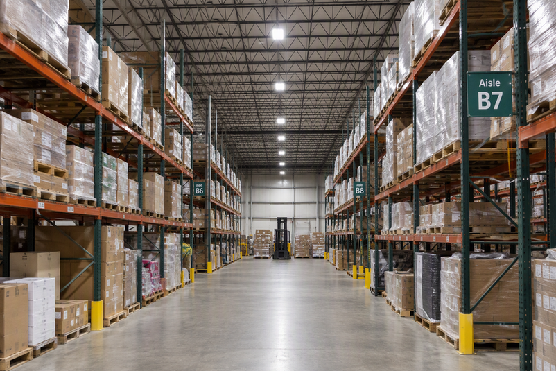 Warehouse aisle beneath exposed roof trusses, with high-bay fixtures at ceiling height