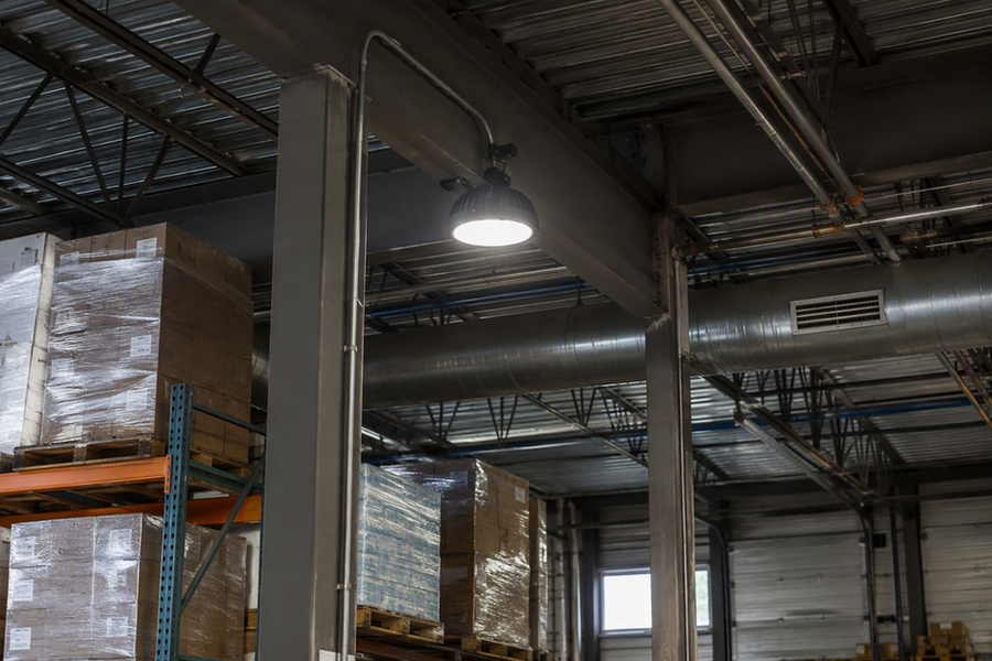 A warehouse aisle beneath a low roof structure, where beams, ducts, and sprinkler lines leave limited room for mounting a high-bay light.