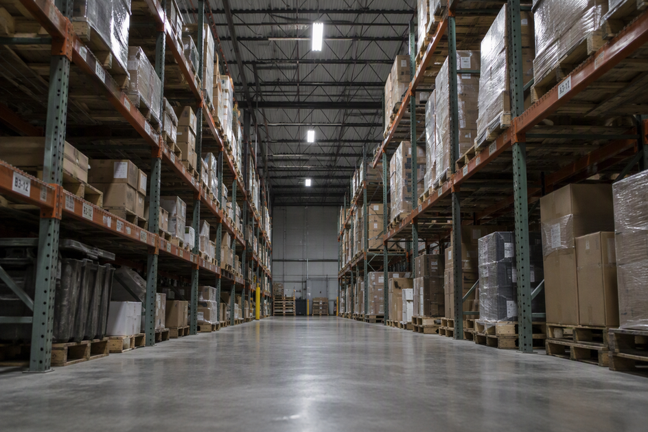 A warehouse rack row sits offset beneath ceiling-mounted linear lights, with shelving between a fixture and the pick face.