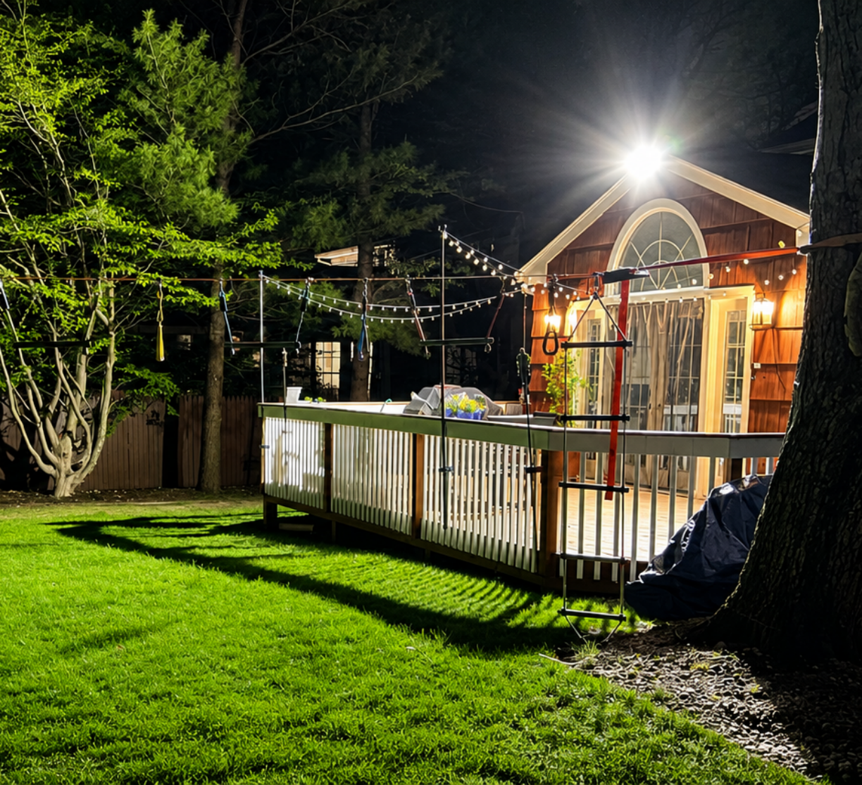 Bright outdoor flood light illuminating a lush green lawn and wooden fence at night, casting clear shadows near a backyard wooden deck.